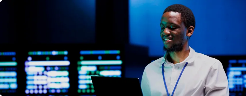 An engineer smiling at a laptop in front of a wall of monitoring dashboards